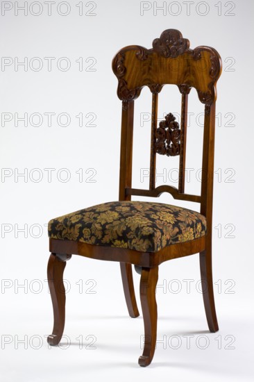 Chair, mahogany chair with a padded seat and a high openwork backrest, with carved decoration. Square legs, S-shaped at the front with volute feet, and arched rear legs. Wavy frame with rounded front corners, with a seat in the form of an overlay cushion. The backrest is framed on the sides by straight stiles extending the rear legs, topped with a board with a symmetrical, wavy-cut outline, emphasized by carved acanthus appliqués. The filling consists of two straight stiles covered with astragalus, topped and fastened together with acanthus leaf arrangements, and a narrow lower stile, arched between the stiles. Acanthus ornament; plant decoration; Louis Philippe style. 1850–1860, Poland, possibly Warsaw. Seating furniture; height 118.7 cm, width 50.6 cm, depth 54.5 cm., A wooden chair with a polished brown finish and carved decorative details on the top of the backrest and the central slat, an upholstered seat covered in a patterned fabric showing shades of black, gold, and muted green...