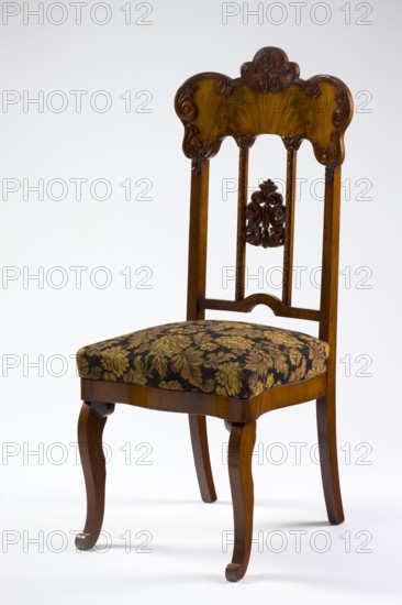 Chair, mahogany chair with a padded seat and a high openwork backrest, with carved decoration; square legs—S-shaped at the front with volute feet—and arched rear legs; wavy frame with rounded front corners and a seat in the form of an overlay cushion; backrest framed on the sides by straight stiles extending the rear legs, topped with a board with a symmetrical wavy-cut outline emphasized by carved acanthus appliqués; the filling has two straight stiles covered with astragalus, topped and fastened together with acanthus leaf arrangements, and a narrow lower stile arched between the stiles; plant decoration, acanthus ornament; Louis Philippe style; dated 1850–1860; origin Poland, possibly Warsaw; seating furniture; height 118.5 cm, width 51 cm, depth 55.7 cm., A wooden chair with a carved top rail and decorative carved central splat, featuring a dark brown polished finish and curving vertical supports, with a cushioned seat upholstered in a patterned fabric showing muted gold and olive ...