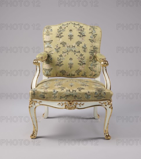 Armchair, Rococo style, floral decoration, cabriole legs, silk fabrics, 18th-century patterned floral fabrics. Upholstered seat, backrest, armrests, carved, white-lacquered and gilded. Cabriole legs, arched into the frame, cut out in a wave-like manner at the bottom; trapezoidal seat with a slightly bulging front frame. Disc-shaped backrest, nearly square, topped with an arch, with slightly concave side rails, straight in line with the sitter's back, slightly tilted with a clearance. Armrests with a volute at the front end, on recessed, slightly S-shaped supports. Edges of all frame elements highlighted with a gilded half-cylinder. Central part of the front frame and upper part of the front legs with carved floral decorations; ends of the armrests and the feet with gilded acanthus leaves. Entire piece reupholstered with cream-colored silk fabric satin-weave background, accented with colored silk and brocaded twill weave with silver thread; decorated with wreaths and alternating vertica...