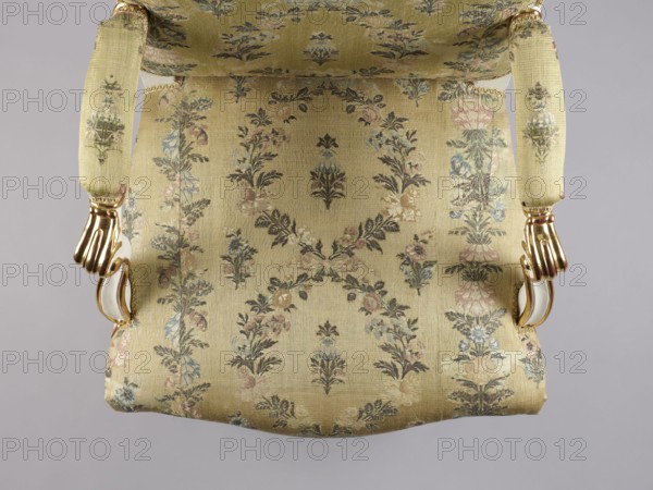 Armchair, Rococo style; floral decoration; cabriole legs; silk fabrics; 18th-century patterned floral fabrics. Upholstered seat, backrest, armrests; carved, white-lacquered and gilded. Cabriole legs arched into the frame, cut out in a wave-like manner at the bottom; trapezoidal seat with a slightly bulging front frame. Disc-shaped backrest, nearly square, topped with an arch, with slightly concave side rails, straight in line with the sitter's back and slightly tilted with a clearance. Armrests with a volute at the front end on recessed, slightly S-shaped supports. Edges of all frame elements highlighted with a gilded half-cylinder. Central part of the front frame and upper front legs with carved floral decorations; ends of armrests and feet with gilded acanthus leaves. Reupholstered with cream-colored silk fabric satin-weave ground, accented with colored silk and brocaded twill weave with silver thread; decorated with wreaths and alternating vertical garlands, with small floral sprigs...