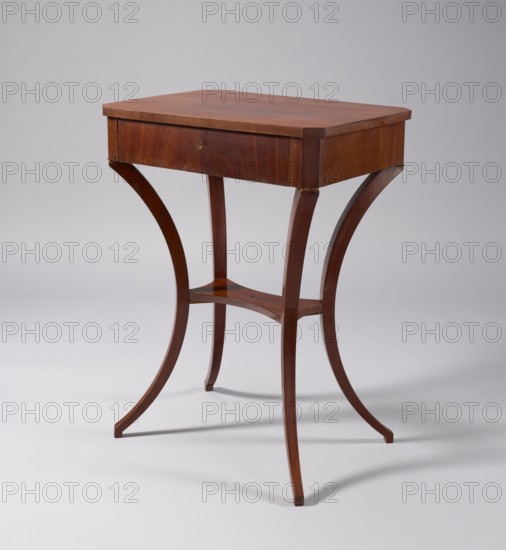 Table, classicism, pre-war collection. TABLE with a rectangular top with chamfered corners, with a drawer in the frame, veneered with mahogany. Supported by four angular outward-curved legs joined mid-height by a shelf with semicircularly indented sides, originally with a finial in the middle. The chamfered corners of the frame and the outer surfaces of the legs at the top decorated with brass rosettes, the drawer lock plate is of a similar form. Mortise lock not preserved. Early 19th century, Poland, Central Europe. Furniture, table; height 78 cm, width 57.8 cm, depth 41.5 cm., A small wooden table with a rectangular top and a shallow drawer, finished in warm brown tones, stands on four curved legs that flare outward and meet at a small curved stretcher; the wood shows variations of reddish-brown and darker brown grain, and the background is a light gray.