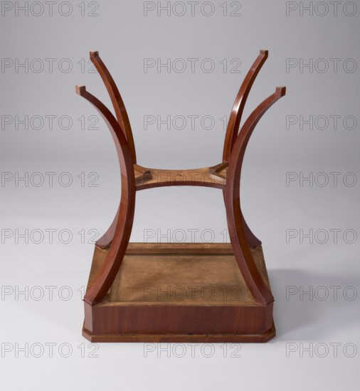 Table, Wereszczakowa, Karolina 1861–1925; classicism; gift; pre-war provenance. TABLE with a rectangular top with chamfered corners, with a drawer in the frame, veneered with mahogany. Supported by four angular outward-curved legs joined mid-height by a shelf with semicircularly indented sides, originally with a finial in the middle. The chamfered corners of the frame and the outer surfaces of the legs at the top decorated with brass rosettes, the drawer lock plate is of a similar form. Mortise lock not preserved. Early 19th century, Poland, Central Europe. Furniture, table. Dimensions: height 78 cm, width 57.8 cm, depth 41.5 cm., The image shows an upside-down wooden stool or small table base with a warm reddish-brown finish, curved legs that arch upward, a rectangular recessed platform at the bottom with a lighter brown interior surface, and a smooth neutral gray background.