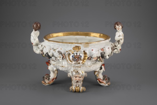 Vase from the swan service, Koenigliche Porzellain Fabrique, Meissen 1710–1763; Eberlein, Johann Friedrich 1695–1749, sculptor; Kaendler, Johann Joachim 1706–1775, co-creator. Soup tureen; oval vase on four molded legs in the form of spits with a molded mask of a bearded man or a volute — a molded dolphin with raised body and tail two, on which the vase is supported. Two handles at the shorter edges in the form of fully modeled naked children — tritons with double decoratively crossed fish tails. Belly is knobbed and modeled with a shell motif in the lower part and a wave band in the upper part, with applied plastic flowers, polychrome-painted shells and seafood, and two oval shields in rocaille-mould cartouches bearing a marital coat of arms supported by heraldic lions with jewels. At the center of the base — a convex gilded shell oval. No cover. Date: 1737–1741. Place of origin: Meissen, Saxony. Vessel. Height 24.0 cm; width 34.0 cm., A decorative ceramic bowl rests against a gray ba...