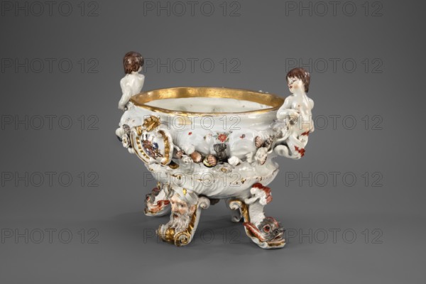 Vase from the Swan service, Koenigliche Porzellain Fabrique, Meissen 1737–1741. Makers: Johann Friedrich Eberlein 1695–1749, Johann Joachim Kaendler 1706–1775. Swan service; soup tureen form, oval vase on four modeled legs in the form of spits with plastic masks of bearded men or volutes—plastic dolphins with raised bodies and tails supporting the vase two. Two handles at the short ends in the form of fully modeled naked children—tritons with double decoratively crossed fish tails. Belly is knobbed and modeled with a shell motif in the lower part and a wave band in the upper part, with applied plastic flowers, polychrome-painted shells and seafood, and two oval shields in rocaille-moulure cartouches bearing a marital coat of arms supported by heraldic lions with jewels. Central underside features a convex gilded shell oval. No cover. Meissen, Saxony. Vessel; height 24.0 cm, width 34.0 cm., A decorative porcelain bowl sits against a gray background, primarily white with gold accents alo...