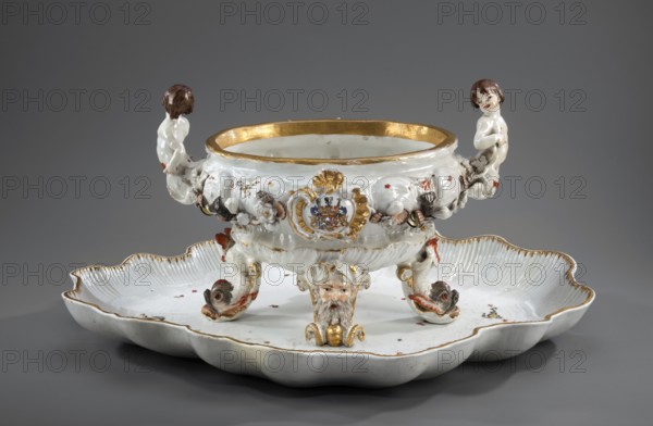 Vase from the swan service, Koenigliche Porzellain Fabrique, Meissen 1737–1741. Authors: Eberlein, Johann Friedrich 1695–1749; Kaendler, Johann Joachim 1706–1775. Oval vase on four applied legs formed as a spit with a plastic mask of a bearded man or a volute — a plastic dolphin with raised body and tail two, supporting the vase. Two handles at the short ends in the form of fully modeled naked children — tritons with double decoratively crossed fish tails. Belly is knopbed and modeled with a shell motif in the lower part and a wave band in the upper part, with applied plastic flowers, polychrome-painted shells and seafood, and two oval shields in rocaille-shell cartouches bearing a marital coat of arms supported by heraldic lions with jewels. Central underside has a convex gilded shell oval. No cover. Meissen, Saxony. Vessel; height 24.0 cm, width 34.0 cm., A decorative porcelain centerpiece sits on a scalloped shallow dish, both predominantly white with gilt edging and accents. The ce...