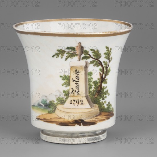 Cup, Korzec Porcelain and Faience Manufactory; 1790–1832. Chalice-shaped cup with stepped foot and curved handle ending in a bird's head. White with a greyish hue, painted in light greens, ochres, and browns, and gilded. Front shows a romantic landscape with ruins and trees and two boulders: the larger inscribed Zaslaw and the smaller dated 1792, topped by a statue of a standing man. Back shows a slender tree with two branches, with shrubs and mosses below. Floral motifs with stylized gold petals on either side of the handle. Inscription and date indicate the site of the victorious Battle of Miropol during the Polish–Russian War following the adoption of the Constitution of May 3rd. Dated ca. 1800–1815. Place of manufacture: Korzec, Volhynian Governorate, Poland. Vessel; height 7.0 cm, width 8.5 cm, depth 7.0 cm., A white porcelain cup with a flared rim and a rounded foot sits against a gray background, the rim edged in a gold color that shows slight wear; the main painted scene on the...