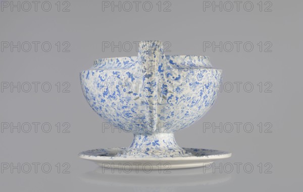 Semi-ovoidal vase on a low pedestal base with sharply broken edges and tapering concave profile. Vertical handles extend slightly beyond the vase width and rise high, modeled as fan-shaped fluted leaves. Decorated with a blue printed wallpaper pattern. Shape based on Wedgwood products see 1817 catalogue, pl.7, fig.146. Maker: Cmielów faience and porcelain factory est. 1804. Prints, wallpaper patterns; unknown provenance. Dated 1840–1860, Poland. Vessel; height 16.0 cm, width 20.0 cm, depth 32.0 cm., A ceramic vessel and matching shallow saucer sit against a pale gray background; the vessel has a rounded bowl shape on a short pedestal foot with a vertical handle-like ridge rising from rim to base, and both pieces are covered in a dense floral pattern in blue on a white ground with areas of light shadow and subtle surface texture visible.