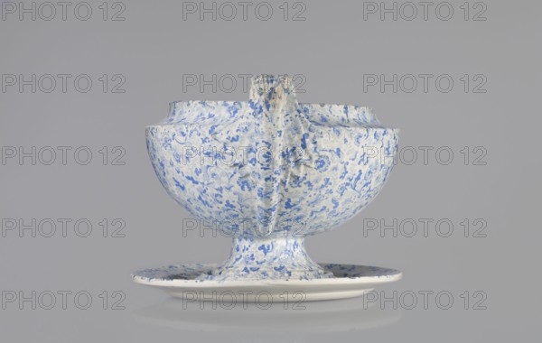 Vase on base. Semi-ovoidal vase on a low pedestal base with sharply broken edges and tapering with a concave line. Vertical handles extend slightly beyond the vase width, raised high in the shape of fan-shaped fluted leaves. Made of faience and porcelain; blue print decoration in a wallpaper pattern. Shape based on Wedgwood's products see 1817 catalogue, pl.7, fig.146. Maker: Cmielów faience and porcelain factory; est. 1804. Date: 1840–1860. Place of origin: Poland. Function: vessel. Dimensions: height 16.0 cm, width 20.0 cm, depth 32.0 cm., A ceramic bowl and matching saucer sit against a plain light gray background, the bowl raised on a short pedestal and featuring a central vertical handle-like ridge; both pieces display a dense floral and foliate pattern in blue on a white base, with areas of the white showing subtle off-white or cream tones and small darker blue accents, and the overall scene includes shades of blue, white, cream, gray, and a touch of darker gray in the shadowing.