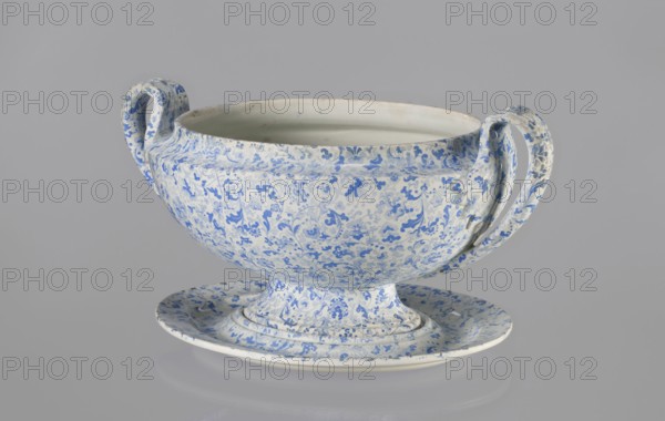 Semi-ovoidal vase on a low pedestal base with sharply broken edges and tapering concave profile. Vertical handles extend slightly beyond the vase width and rise high as fan-shaped fluted leaves. Decorated with a blue print in a wallpaper pattern. Shape based on Wedgwood products see 1817 catalogue, pl.7, fig.146. Made by Cmielów faience and porcelain factory est. 1804. Prints/wallpaper patterns, unknown provenance. Dated 1840–1860, Poland. Vessel; height 16.0 cm, width 20.0 cm, depth 32.0 cm., A ceramic bowl with a pedestal and attached saucer is shown against a plain background; the bowl has two large looped handles and is covered in an intricate floral and foliate pattern rendered in blue on a white ground, with the interior of the bowl appearing mostly white with slight blue mottling, and the background and reflected surface showing shades of gray.