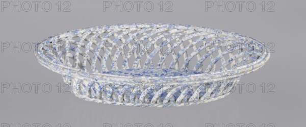 Openwork basket, Cmielów faience and porcelain factory founded 1804. Openwork basket with an oval, mandorla-like cross-section and parted walls formed by two layers of rollers rotating diagonally in opposite directions. Top enclosed in a band of three rollers; bottom finished in a braided-rope form. Interior bottom fitted with an oval plate. Surface decorated with a blue wallpaper pattern. Abrasions to the glaze and slight chipping on the rim. Dated 1840–1860. Poland; Cmielów. Ceramic vessel. Height 6.5 cm; length 30.5 cm; width 18.5 cm., A shallow oval woven basket with an open lattice structure made from twisted strips, covered in a patterned surface showing white and blue as the dominant colors with small areas of beige and faint gray; the weave creates curved diagonal openings around the sides and a flat base with a floral-like motif, all set against a plain light gray background.