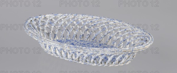 Openwork basket. Faience and porcelain factory, Cmielów est. 1804. Openwork, basket with an oval cross-section similar to a mandorla with parted walls, formed by two layers of rollers rotating diagonally in two opposite directions. Top enclosed in a band of three rollers; bottom finished in a kind of braided rope. Interior bottom plastered with an oval plate. Surface decorated with a blue wallpaper pattern. Abrasions to the glaze and slight chipping on the edge of the walls. Dated 1840–1860. Poland. Cmielów. Vessel. Height 6.5 cm; length 30.5 cm; width 18.5 cm., A woven oval basket with an open lattice structure made of curved interlacing rods, featuring a patterned surface in white and blue with touches of light beige, containing a patterned cushion that matches the basket's white and blue color scheme; the background is a uniform light gray.