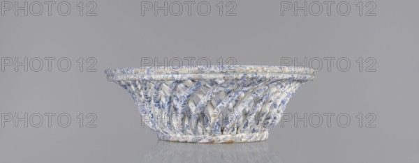 Openwork basket, faience and porcelain, Cmielów factory est. 1804. Openwork, wallpaper-pattern surface. Basket with an oval mandorla-like cross-section and parted walls formed by two layers of diagonally rotating rollers in opposite directions; top enclosed in a band of three rollers, bottom finished in a braided-rope form. Inner bottom plastered with an oval plate. Surface decorated with a blue wallpaper pattern. Abrasions to glaze and slight chipping on wall edge. 1840–1860, Poland. Vessel; height 6.5 cm, length 30.5 cm, width 18.5 cm., A shallow woven basket with an open lattice of twisted bands displayed against a plain gray background, the basket showing a white base color with a delicate blue floral pattern across its surface and small areas of brownish discoloration, the rim slightly thicker and decorated with the same white and blue motif, the basket casting a soft reflection on the surface beneath it.