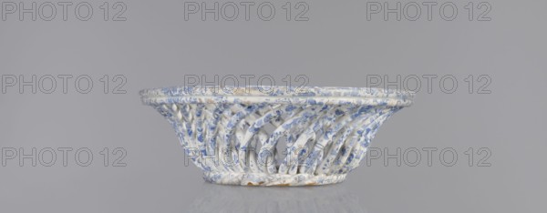 Openwork basket, Cmielów faience and porcelain factory est. 1804. Openwork, oval cross-section similar to a mandorla with parted walls formed by two layers of rollers rotating diagonally in opposite directions. Top enclosed in a band of three rollers; bottom finished in a kind of braided rope. Interior bottom plastered with an oval plate. Surface decorated with a blue wallpaper pattern. Abrasions to the glaze and slight chipping on the edge of the walls. 1840–1860, Poland. Vessel; height 6.5 cm, length 30.5 cm, width 18.5 cm., A shallow woven bowl with a lattice-like shape made of twisting curved strips, primarily white with blue floral and decorative patterns, showing some small areas of brownish discoloration or wear, set against a plain light gray background and reflected faintly on a glossy surface.