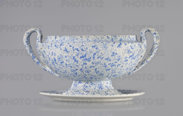 Vase, Cmielów faience and porcelain factory; 1804–, wallpaper pattern, orchids, plant and floral decoration, unknown provenance. Small vase with a convex belly supported by a small foot. Decoration in blue print with a wallpaper pattern. Vertical, fan-shaped handles, slender in a fantastic leaf shape with a line gently diverging from the belly and curling high towards the rim, surrounding the upper part of the vase with a fluted leaf. 1840–1860, Poland, Cmielów. Vessel; height 16 cm, length 32 cm, width 20 cm., A ceramic two-handled bowl with a matching underplate, both covered in an intricate floral and vine pattern in shades of blue on a white background, with sweeping curved handles on either side and a rounded foot supporting the bowl; the surface shows subtle variations in blue intensity and some small areas of wear, and the objects sit against a smooth gray background with a faint reflection visible beneath the plate.