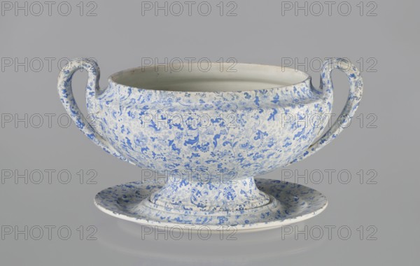 Vase, Cmielów faience and porcelain factory; 1804-, wallpaper patterns, orchids, plant decoration, floral decoration, unknown provenance. A small vase with a convex belly supported by a small foot. Decoration in blue print with a wallpaper pattern. Vertical, fan-shaped handles, slender in a fantastic leaf shape with a line gently diverging from the belly and curling high towards the rim, surrounding the upper part of the vase with a fluted leaf. 1840–1860, Poland, Cmielów; vessel; height 16 cm, length 32 cm, width 20 cm., A ceramic two-handled bowl on a matching shallow saucer, both covered in an intricate floral and vine pattern in shades of blue on a white background; the bowl has a rounded body with a wide open rim and curved handles rising from either side, and the saucer mirrors the blue-and-white motif with a raised central pedestal supporting the bowl; visible colors include blue, white, and subtle off-white or cream tones, set against a neutral light gray backdrop.