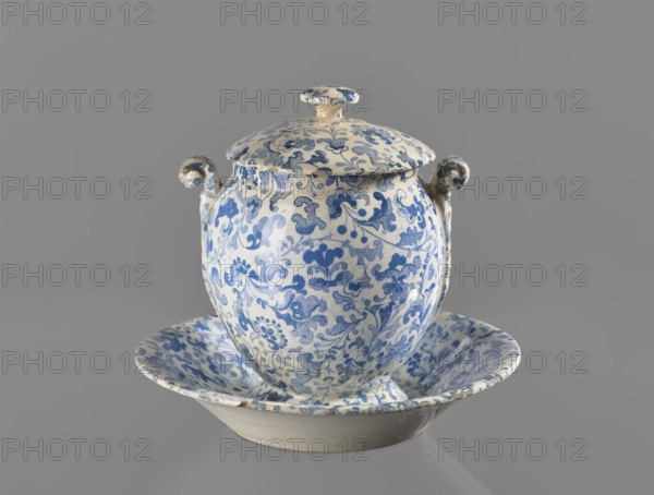 Mustard dish with lid, Cmielów faience and porcelain factory; 1804-, floral decorations. Mustard dish with an ovoid bowl, volute handles at the edge, combined with a plate-shaped base. Slightly domed lid with a mushroom handle and a cutout for a spoon. Decoration with a blue print—wallpaper pattern with a spirally twisted tendril with fancifully twisted leaves and flowers of various sizes in the orchid type. 1840–1860. Poland; Cmielów. Vessel. Height 10.5 cm; width 12.0 cm., An ornate lidded ceramic vessel with a rounded body and two small curled handles sits centered on a shallow matching dish; both pieces display an all-over intricate floral and foliate pattern in blue on a pale cream background, with the lid topped by a small knob also patterned in blue and cream, and the dish showing a flared rim with the same blue floral decoration, all set against a solid neutral gray backdrop.