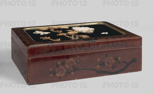 Box, Rectangular box with a put-on lid, decorated with red lacquer painted with a pattern of blooming cherry blossoms. On the lid, a slightly concave black field with rounded corners, with a narrow gilded frame, inlaid carved decoration of hydrangea branches, a bird, and a butterfly. The branches and leaves are dyed green. Inside, four small boxes with lids, decorated with red lacquer painted with a pattern of cherry blossoms and branches. 19th/20th century, Japan. Container. Height 8 cm, width 26 cm, depth 19 cm., A rectangular lacquered box with a hinged lid sits on a neutral surface, showing a glossy reddish-brown exterior decorated with dark brown and black floral and branch motifs; the top panel features a recessed black inset bordered by a thin metallic rim and adorned with several raised carved elements in light tan and beige forming flower and leaf shapes on twig-like brown pieces; the sides of the box display painted blossom designs in muted gold and dark greenish-black; visib...