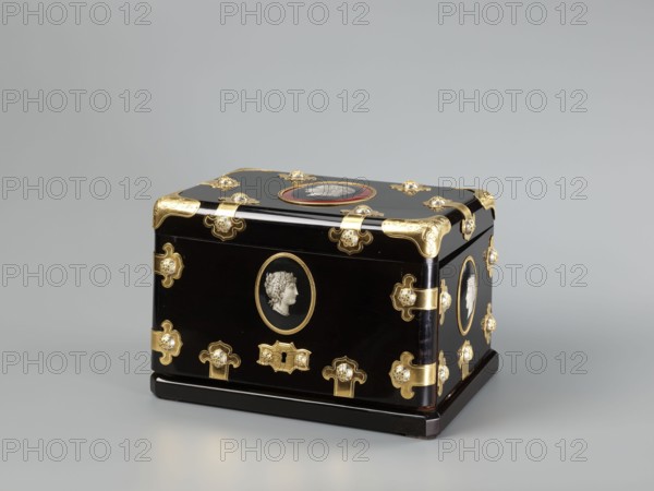 Cigar box, cigars, cameos, antiquing style, smoking tobacco, female busts, male busts, 3rd quarter of the 19th century, France, container; height 18.7 cm, width 28.5 cm, depth 20 cm, A small rectangular box sits on a neutral gray surface against a pale gray background, finished in a glossy black with several decorative gold metal fittings at the corners, edges, and front, and with small round clear or white jewel-like accents set into some of the fittings; oval portrait medallions with pale relief profiles appear on the front, top, and side surfaces, each set within gold or colored borders; the box rests on a slightly raised black base and shows contrasts of black, gold, pale off-white in the relief portraits, and a touch of red around the top medallion.