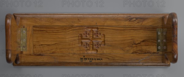 Bookstand, origin unknown, Hebrew, Jerusalem Israel/Palestine; rectangular foldable bookstand with two movable sides, decorated with three recessed Jerusalem Crosses. Under the central cross an ink Hebrew inscription reads ??????? Yerushalayim. Main part has profiled edges and can widen by shifting the central part in a groove to the right. Movable sides have a heart-shaped outline, unfold on brass hinges to an almost vertical position with slight inclination, and are decorated with crosses on the outer surface. 19th–20th century; hardware; height 12.6 cm, width 37.3 cm, depth 2.9 cm., A rectangular wooden tray viewed from above with rounded handles on each short end and a recessed central panel showing natural wood grain patterns in shades of brown and tan; the panel has a carved symmetrical cross-like motif in the center and two brass hinge plates set into the interior edges near the handles, with visible metal screws; the wood shows darker brown streaks and knots against a lighter b...