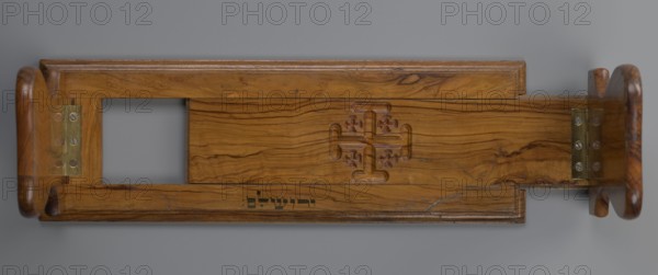 Bookstand, origin unknown, Hebrew; rectangular wooden bookstand with two foldable sides, decorated with three recessed Jerusalem crosses. Under the central cross is an ink inscription in Hebrew: ??????? Yerushalayim. Main part has profiled edges and can be widened by shifting the central part embedded in a groove to the right. Movable sides have a heart-shaped outline; when unfolded on brass hinges they sit almost vertically with a slight inclination and are decorated with crosses on the outer surface. 19th–20th century; possible origin Jerusalem; material: hardware; height 12.6 cm, width 37.3 cm, depth 2.9 cm., A horizontal wooden object with a warm medium-brown grain and darker brown streaks, shown against a neutral gray background, featuring a carved symmetrical cross-like motif with rounded ends in the center panel, a rectangular cutout on the left side, metal hinge plates with a brass tone and visible fasteners near both ends, rounded end pieces with smooth contoured profiles, and...