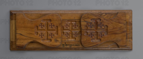 Bookstand, origin unknown, Hebrew, Jerusalem Israel/Palestine; rectangular bookstand with two foldable sides, decorated with three recessed Jerusalem Crosses. Under the central cross is an ink inscription in Hebrew: ??????? Yerushalayim. Main part has profiled edges and can widen by shifting the central part embedded in a groove to the right. Movable sides have a heart-shaped outline; when unfolded on brass hinges they sit nearly vertical with a slight inclination and are decorated with crosses on the outer surface. 19th/20th century; hardware; height 12.6 cm, width 37.3 cm, depth 2.9 cm., A rectangular wooden panel with a warm brown color and visible wood grain, featuring a carved design of a group of rounded rectangular shapes with raised cross-like motifs repeated across them; the wood shows darker brown veining and a few blackish streaks, with carved grooves and recessed areas creating shadows of deeper brown; the panel has a framed border in a similar brown tone and the background...