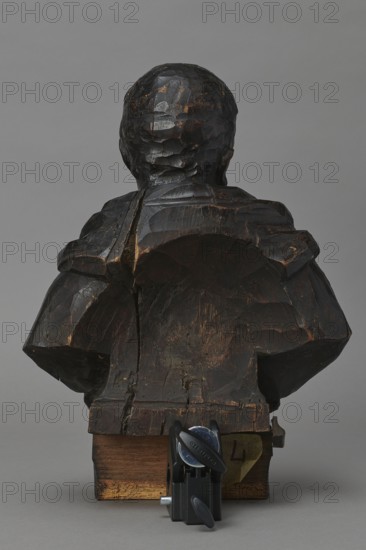 Bed finial from the Hospital of St. John of God in Warsaw. Maker: Redler, Jan Chryzostom 1704–1779, woodcarver bust; Designer: Fontana, Jakub 1710–1773 design. Hospital bed finial in the form of a pediment with volutes on the sides and a corbel in the middle supporting a female bust; fastened with two pegs to the front section of the cornice crowning the canopy. Figure dressed in a tunic, head turned to the right, hair pinned up. Outline of the finial highlighted with a black ribbon stucco inlay; corbel with slats. On the corbel, an oval hole containing a wooden rotating medallion with inlaid letters on obverse and reverse medallion missing; above it an inlaid number illegible. The runners originally had garlands; the volutes had small brackets with turned, cup-shaped pinnacles. Date: ca. 1757–1760, Warsaw. Fragment of furniture. Dimensions: height 75 cm, width 95.5 cm, depth 14 cm., The image shows the rear view of a dark brown carved wooden bust mounted on a lighter brown wooden base...