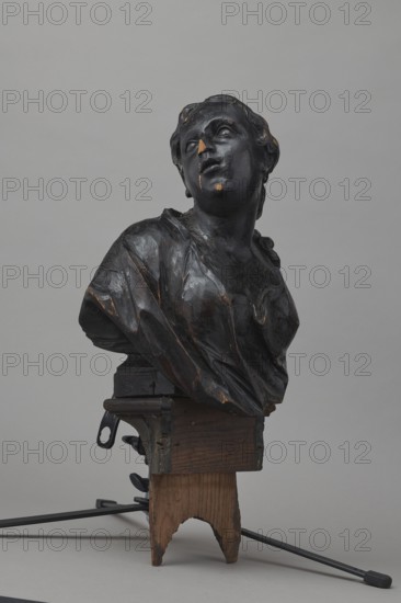 Bed finial, ca. 1757–1760. Baroque wooden bed finial in the form of a pediment with volutes on the sides and a corbel in the middle supporting the bust of a young woman; fastened with two pins to the front section of the cornice crowning the canopy. The outline of the finial and the corbel are highlighted with a black ribbon stucco inlay. The corbel has an oval opening filled with a wooden, rotating medallion with inlaid letters on the obverse and reverse missing. The runners feature garlands made of ribbons and bay leaves; the volutes feature small brackets with turned, cup-shaped pinnacles. Attributed designers and makers: Jakub Fontana 1710–1773, designer; Jan Chryzostom Redler 1704–1779, woodcarver. Origin: Warsaw. Function: furniture fragment hospital bed finial. Dimensions: height 66 cm, width 93 cm, depth 16 cm., A dark bust of a person with a tilted head and closed eyes sits on a wooden support; the bust has a glossy black surface with worn areas showing a warm brown or golden ...
