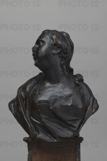 Bed finial in the form of a pediment with volutes on the sides and a corbel in the middle supporting the bust of a young woman; fastened with two pins to the front section of the cornice crowning the canopy. The outline of the finial and the corbel are highlighted with a black ribbon stucco inlay. The corbel has an oval opening filled with a wooden, rotating medallion with inlaid letters on the obverse and reverse missing. The runners feature garlands made of ribbons and bay leaves; the volutes feature small brackets with turned, cup-shaped pinnacles. Female busts; baroque style. Made ca. 1757–1760, Warsaw. Makers: Jakub Fontana 1710–1773 designer, co-creator, Jan Chryzostom Redler 1704–1779 woodcarver, author. Furniture fragment; height 66 cm, width 93 cm, depth 16 cm., A dark wooden bust of a person turned to the side, showing a head with carved hair and a neck flowing into draped clothing; the surface has worn spots revealing lighter brown beneath the dark finish, and the piece rest...