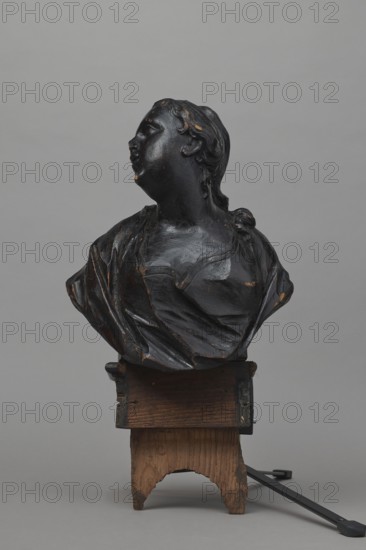Bed finial, ca. 1757–1760. Baroque wooden hospital bed finial in the form of a pediment with volutes at the sides and a central corbel supporting the bust of a young woman; fastened with two pins to the front section of the cornice crowning the canopy. Outline of the finial and corbel highlighted with a black ribbon stucco inlay. Corbel has an oval opening that once contained a wooden rotating medallion with inlaid letters on obverse and reverse missing. Runners feature garlands of ribbons and bay leaves; volutes have small brackets with turned, cup-shaped pinnacles. Makers: Jakub Fontana 1710–1773, designer; Jan Chryzostom Redler 1704–1779, woodcarver. Origin: Hospital of St. John of God, Warsaw. Furniture fragment. Dimensions: height 66 cm, width 93 cm, depth 16 cm., A dark-toned bust of a person viewed in three-quarter profile, with the head turned slightly upward and to the side, showing textured hair and a smooth face with subtle wear spots; the clothing is draped with visible fol...