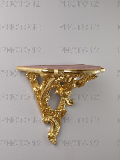 Hanging console, acanthus ornament, floral ornament, Rococo style, ca. mid-18th century, Central Europe, piece of furniture; H. 37.3 cm, W. 41.5 cm, D. 26 cm, A wall-mounted decorative shelf with ornate carved scrollwork in a shiny gold finish supporting a flat top that appears reddish-brown, mounted against a smooth light gray wall; the gold carving features flowing leaf and floral motifs and curved supports beneath the top.