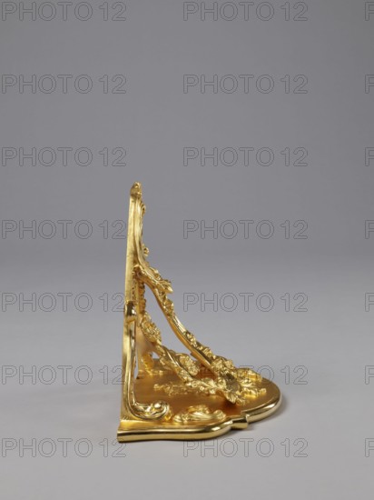 Hanging console, acanthus ornament, floral ornament, rococo style, ca. mid-18th century, Central Europe, piece of furniture; height 37.3 cm, width 41.5 cm, depth 26 cm, The image shows an ornate decorative bracket or support placed on a smooth surface against a plain background, rendered in a shiny gold color with a polished finish; the piece features flowing, carved motifs with curved lines, leaf-like forms, and rounded scroll details, and there are highlights and subtle shadows accentuating its three-dimensional sculpted surfaces against a neutral gray backdrop.