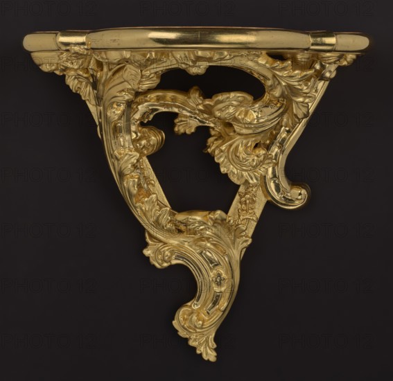 Hanging console, acanthus ornament, floral ornament, Rococo style, ca. mid-18th century, Central Europe, piece of furniture, height 37.3 cm, width 41.5 cm, depth 26 cm, A decorative wall bracket with ornate carved scrolling foliage and acanthus motifs finished in a shiny gold color is shown against a dark background that appears black. The bracket has a curved triangular form with flowing leafy details, rounded molding along the top edge, and smooth reflective surfaces where the gold catches light. The carved areas display layered leaves and volutes, creating pockets of shadow and bright highlights on the metallic gold finish. The overall color palette is gold and black.