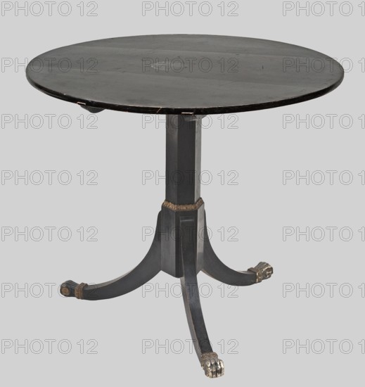 Table, provenance, animal paws; Empire style; cymatium ornament. Round tilting top supported by a hexagonal shaft forked into a tripod, lacquered black. Arched legs topped with gilded and black-painted feet in the shape of animal paws. Shaft widened in the lower section, separated from the upper section by a gilded strip with a lesene cymatium. Top reinforced from below with two slatted pins that are part of the hinge, mounted on a block crowning the stem. Steel latch, mortise-and-apply, with a steel catch mounted on the side of the top. Dated 1810–1825, Central Europe. Piece of furniture; height 74 cm, diameter 85 cm., The image shows a round wooden table with a dark surface and a single central pedestal base that splits into a group of curved legs ending in carved feet; the table and legs are primarily black with areas of worn lighter wood and small patches of gold or brass-colored detailing, set against a light gray background.