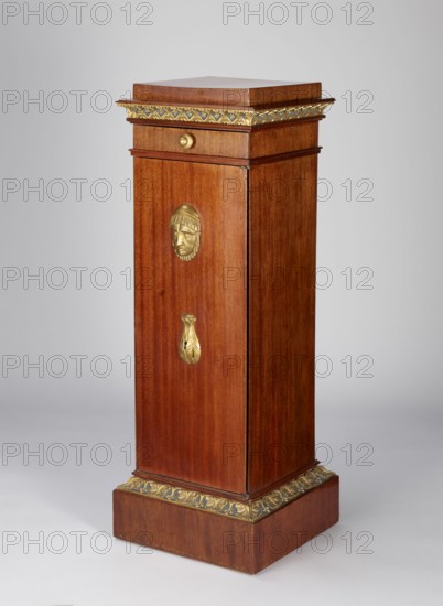 Pedestal cabinet, single-leaf with a drawer at the top, supported by a prominent plinth; veneered with mahogany, with profiled, carved, gilded and painted cornices and applied decoration on the doors. The edges of the door leaf and drawer faces and the corresponding front edges of the walls are diagonally cut. The molding around the plinth is decorated with a dark green and gilded leaf ornament, the cornice under the top is decorated with rosettes set in a rhomboidal grid. The upper section of the door features a bas-relief woman's head, below it a lotus flower with a keyhole; on the drawer face is a gilded wooden handle in the shape of a mushroom. Originally four shelves inside. The drawer bottom is recessed from the back. A single-bolt, self-locking steel mortise-and-apply lock. Brass pintle hinges. Empire style, first quarter of the 19th century. Possible origin: Poland or Austria. Height 97.2 cm, width 33.5 cm, depth 35.2 cm., A tall rectangular wooden pedestal with a warm reddish-...