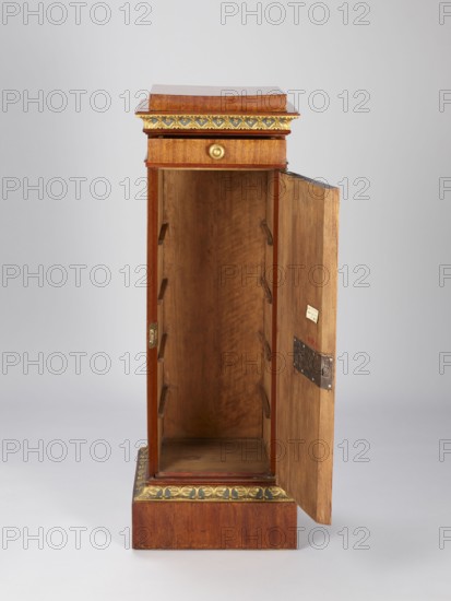 Pedestal cabinet, single-leaf with a drawer at the top, supported by a prominent plinth; veneered with mahogany, with profiled, carved, gilded and painted cornices and applied decoration on the doors. The edges of the door leaf and drawer faces and the corresponding front edges of the walls are diagonally cut. The molding around the plinth is decorated with a dark green and gilded leaf ornament; the cornice under the top is decorated with rosettes set in a rhomboidal grid. The upper section of the door features a bas-relief woman's head; below it a lotus flower surrounding the keyhole; on the drawer face is a gilded wooden handle in the shape of a mushroom. Originally four shelves inside. The drawer bottom is recessed from the back. A single-bolt, self-locking steel mortise-and-apply lock. Brass pintle hinges. Empire style, first quarter of the 19th century; origin possibly Poland or Austria. Materials: mahogany veneer, gilding, painted decoration. Height 97.2 cm, width 33.5 cm, depth ...