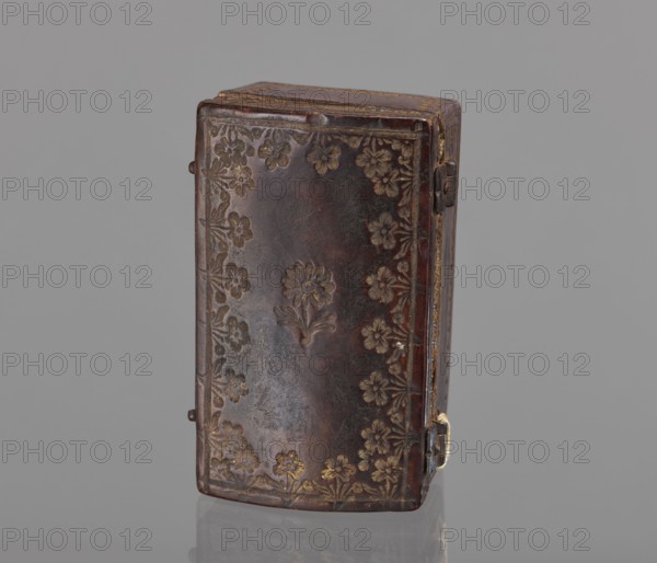 Case for a stamp, bookbinding, embossing; leather case shaped like a chest slightly widened at the top, with an eternal hinge slightly convex, covered with leather with gold-embossed decoration stripes and zigzag lines at the edges; on the lid, flowers with stems at the edge and one in the center. Interior lined with pink satin. 2nd half of the 18th century, Poland . Case; height 3.5 cm, width 7.8 cm., A rectangular box with rounded edges standing upright, covered in dark brown material with a worn, slightly glossy surface and a pattern of embossed floral motifs around the edges and a central flower design; the floral embossing shows lighter brown and muted gold highlights. The box has a visible hinge and small metal clasp on one side with a hint of dull metallic color, and the top edge shows lighter brown wear and a thin gold trim. The background is a uniform mid-gray, and the box sits on a reflective surface that creates a faint mirror of its lower edge.