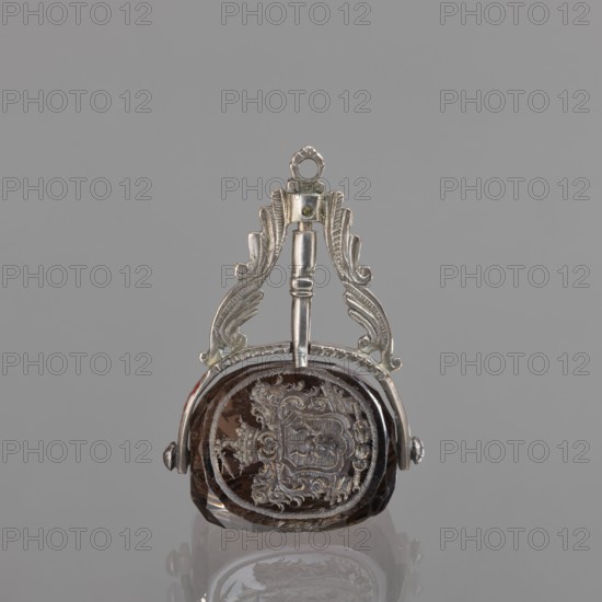 Seal stamp with the Pomian coat of arms; coats of arms; Pomian coat of arms; private seals; coat of arms seals; Axe coat of arms. Seal stamp made of smoke crystal, three-sided with three Polish coats of arms, silver binding with a volute motif. On individual fields: the Bawól head coat of arms; the Pomian coat of arms, a five-field coat of arms: inside Pomian, in the upper two Toxe, an unrecognized crowned rooster, in the lower ones: Rolbiecki and an unrecognized coat of arms. All under noble crowns, with jewels; five-field surrounded by war trophies. 3rd quarter of the 18th century, Poland, stamp; height 5.2 cm, width 3.5 cm., A small ornate pendant-like object with a metallic silver top piece featuring symmetrical scrollwork and a loop for hanging, attached to a faceted dark brown gemstone or glass piece with an engraved decorative design visible on its surface; the metal is silver in color and the stone is dark brown with lighter brown highlights, set against a neutral gray backgrou...