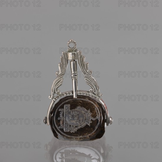 Seal stamp with the Pomian coat of arms; coats of arms; Pomian coat of arms; private seals; coat of arms seals; Axe coat of arms. Seal stamp made of smoke crystal, three-sided with three Polish coats of arms; silver binding with a volute motif. On individual fields: the Bawól head coat of arms; the Pomian coat of arms; a five-field coat of arms: inside Pomian, in the upper two Toxe, an unrecognized crowned rooster, in the lower ones: Rolbiecki and an unrecognized coat of arms. All under noble crowns, with jewels; the five-field surrounded by war trophies. 3rd quarter of the 18th century; Poland, stamp; height 5.2 cm, width 3.5 cm., A small ornamental object with a metallic frame holding a dark translucent stone, shown against a uniform gray background; the metal appears silver in color with decorative curved elements and a loop at the top, and the stone is a dark brown to amber color with visible internal carved or etched shapes; the object casts a faint reflection on the glossy surfac...
