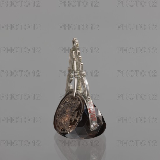 Seal stamp with the Pomian coat of arms; coats of arms; Pomian coat of arms; private seals; coat of arms seals; Axe coat of arms. Seal stamp made of smoke crystal, three-sided with three Polish coats of arms, silver binding with a volute motif. On individual fields: the Bawól buffalo head coat of arms; the Pomian coat of arms; a five-field coat of arms: inside Pomian, in the upper two Toxe, an unrecognized crowned rooster, in the lower ones Rolbiecki and an unrecognized charge. All under noble crowns, with jewels; the five-field surrounded by war trophies. 3rd quarter of the 18th century, Poland, stamp; height 5.2 cm, width 3.5 cm., A small decorative object sits on a reflective surface against a gray background, featuring a faceted dark stone base in deep black and dark brown tones supporting a metallic structure in silver color with an ornate, arched shape; attached to the metal is a round, flat medallion with intricate raised patterns in aged bronze and brown shades, and there are t...