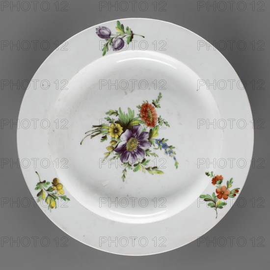 Round platter, Korzec Porcelain and Faience Manufactory 1790–1832. Shallow plate with a wide, flat collar. White porcelain painted with floral motifs: collar has three sprigs with flowers in yellow, purple, and dark vermillion; the mirror shows a large bouquet with a purple anemone and a dark vermillion aster. Muted, cool palette — lemon yellow, dark purple, brown cinnabar — complemented by greens malachite and light yellow. Circa 1810–1815. Origin: Korzec, Volhynian Governorate, Poland. Vessel; height 5.7 cm, diameter 35 cm., A round white ceramic plate with a shallow well, shown against a gray background, decorated with a central floral spray featuring a large purple flower with a yellow center, surrounded by orange, yellow, and small blue blossoms and green leaves, and with additional small floral sprigs near the rim in purple and green, yellow and green, and orange and green; the plate shows subtle surface marks and light reflections.