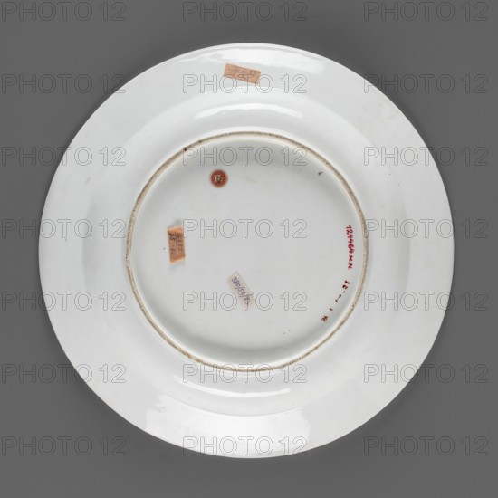 Round platter, Korzec Porcelain and Faience Manufactory; 1790–1832. A shallow plate with a wide, flat collar. White porcelain painted with floral motifs: on the collar three sprigs with flowers in yellow, purple, and dark vermilion; in the mirror a large bouquet with a purple anemone and a dark vermilion aster. Muted tones, cool palette: lemon yellow, dark purple and brown cinnabar complemented by green with malachite and light yellow accents. Circa 1810–1815. Poland, Volhynian Governorate, Korzec. Vessel. Height 5.7 cm; diameter 35 cm., A round white ceramic plate shown from the back on a dark gray background, with a recessed central footring and a shallow well, showing glossy highlights and some areas of light wear and faint smudging; a small round sticker with red and gold, a few rectangular paper stickers in shades of tan and pale blue, and a short red mark near the inner rim are visible, along with subtle beige discoloration around the footrim and small dark specks on the surface.