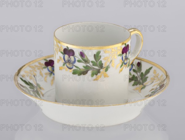Cup and saucer, Korzec Porcelain and Faience Manufactory, 1790–1832. Colorfully painted with a pattern of golden twigs and pansies. 1800–1820. Poland. Vessel., A white porcelain cup sits inside a matching saucer, both decorated with painted floral motifs and gilded rims; the cup shows a band of painted flowers with purple, blue, and green hues accented by gold leaf-like detailing, and a gold-painted handle, while the saucer repeats the floral and gold decoration around its inner edge against the white background, with visible shades of white, gold, purple, blue, and green.