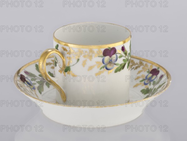 Cup and saucer, Korzec Porcelain and Faience Manufactory 1790–1832, colorfully painted with a pattern of golden twigs and pansies, 1800–1820, Poland, vessel, A white porcelain cup with a matching shallow saucer, both decorated with hand-painted floral motifs in purple, blue, green, and yellow, and trimmed with gold along rims and on a curved gold handle; the cup shows a band of gold-accented foliage and painted flowers, while the saucer displays similar floral decorations around its inner edge against a pale neutral background.