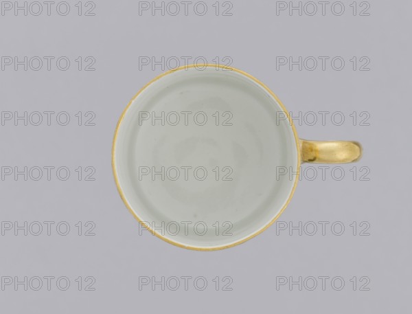 Cup with pansies, Korzec Porcelain and Faience Manufactory 1790–1832. Cylindrical cup litron. Around the upper part of the bowl under the rim, a trail of gilded twigs and colorful pansies; gilded rim. Loop handle, gilded. 1805–1821. Korzec, Volhynian Governorate. Vessel. Height 6.1 cm; width 8.8 cm; diameter 6.5 cm., A top-down view of a round cup with a pale interior and a narrow gold rim, showing a gold handle extending to the right; the background is a uniform light gray and the visible colors include white or pale off-white, gold, and gray.