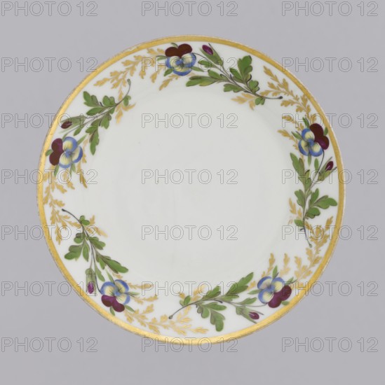 Saucer with floral decoration, Korzec Porcelain and Faience Manufactory; 1790–1832. Conical saucer with a trail of gilded leaves and colorful pansies around the collar; gold border around the edge. Pansies, floral decorations. 1805–1821. Korzec, Volhynian Governorate. Vessel. Height 2.7 cm; diameter 13.5 cm., A round white plate with a gilded rim and a decorative border of green leafy sprigs and golden fern-like fronds, punctuated by a few purple and blue pansy-like flowers and small purple buds, set against a pale gray background.