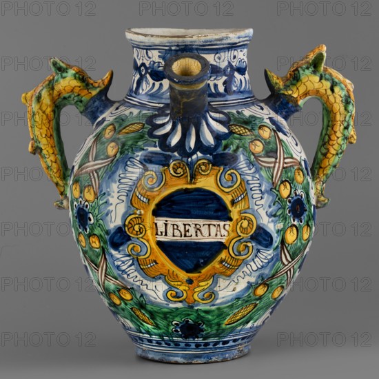 Pharmacy jug, Montelupo ceramics center; ca. 1400–, role classification of maker, pottery inscriptions — dragons, monograms, cartouches, plant decorations, jugs. A two-handled, ovoid pharmacy vase with a cylindrical neck, gently flaring at the rim. On the belly is a tubular spout, and on the sides are two handles in the form of fanciful dolphins dragons?. On the front is an oval cartouche with the inscription LIBERTAS framed by a massive laurel garland. The remaining background is grey and white, painted in two tones of cobalt, with a densely coiled twig with tripartite leaves and flowers a foglie. The decoration is dominated by cobalt blue in varying degrees of saturation, yellow, and green with accents of ochre and purple. Around the neck is a wavy twirl with birds. Below the handles is the letter B. The vase is an interesting example of Montelupo production, specializing in pharmacy vessels with leaf decoration inspired by Venetian ornamentation. Circa 1575, Italy, Montelupo, vessel...
