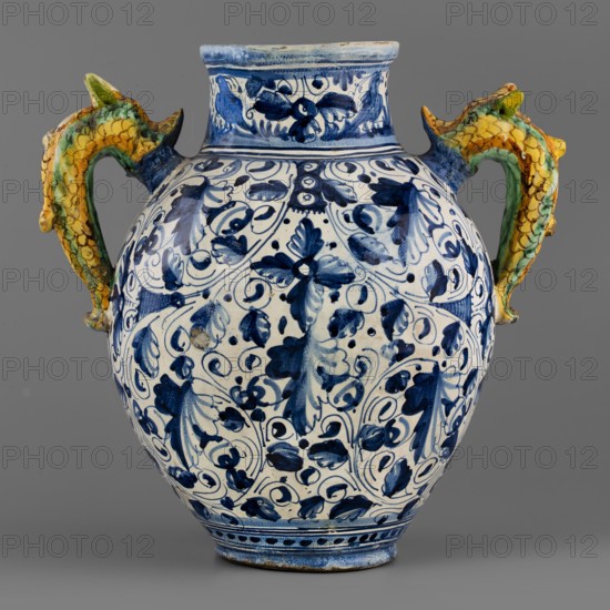 A two-handled, ovoid pharmacy vase with a cylindrical neck, gently flaring at the rim. On the belly is a tubular spout, and on the sides are two handles in the form of fanciful dolphins dragons?. On the front is an oval cartouche with the inscription LIBERTAS framed by a massive laurel garland. The remaining background is grey and white, painted in two tones of cobalt, with a densely coiled twig with tripartite leaves and flowers a foglie. The decoration is dominated by cobalt blue in varying degrees of saturation, yellow, and green with accents of ochre and purple. Around the neck is a wavy twirl with birds. Below the handles is the letter B. The vase is an example of Montelupo production, specializing in pharmacy vessels with leaf decoration inspired by Venetian ornamentation. Circa 1575, Montelupo, Italy; ceramic vessel, height 36 cm, width 29 cm., A ceramic vase with a rounded body and a short neck sits against a gray background, decorated overall with blue floral and foliate brush...