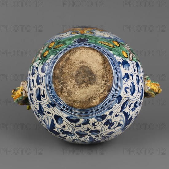 Pharmacy jug, Montelupo ceramics center; ca. 1400–, role classification of maker, pottery inscriptions — dragons, monograms, cartouches, plant decorations, jugs. A two-handled, ovoid pharmacy vase with a cylindrical neck, gently flaring at the rim. On the belly is a tubular spout, and on the sides are two handles in the form of fanciful dolphins dragons?. On the front is an oval cartouche with the inscription LIBERTAS framed by a massive laurel garland. The remaining background is grey and white, painted in two tones of cobalt, with a densely coiled twig with tripartite leaves and flowers a foglie. The decoration is dominated by cobalt blue in varying degrees of saturation, yellow, and green with accents of ochre and purple. Around the neck is a wavy twirl with birds. Below the handles is the letter B. The vase is an interesting example of Montelupo production, specializing in pharmacy vessels with leaf decoration inspired by Venetian ornamentation. Circa 1575, Italy, Montelupo; vessel...