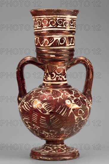 Amphora-type vase, Manises ceramic center; ca. 1400- Maker/ workshop unknown. Two-handled vase with spherical body on a spool base, tall pot-like spout and neck. Painted in red mirror paint with ornamental motifs in a Middle Eastern style; on both sides of the body a bird motif among leaves and stylized plant branches. Neck and spout decorated with a double trail with a volute ornament. Dark cream glaze; glazed underside. 17th century, Spain, Valencia, Manises. Vessel. Height 26.5 cm; width 13.0 cm., An earthenware jug with a rounded body, a narrow neck that flares at the rim, a footed base, and two curved handles; the surface shows decorative painted motifs in a reddish-brown color against a cream background, with swirling and curved patterns covering the neck, shoulder, and body, and bands of ornamentation around the rim and foot; the jug has a glossy finish with some worn areas and small chips, and the visible colors are reddish-brown, cream, and small patches of darker brown.