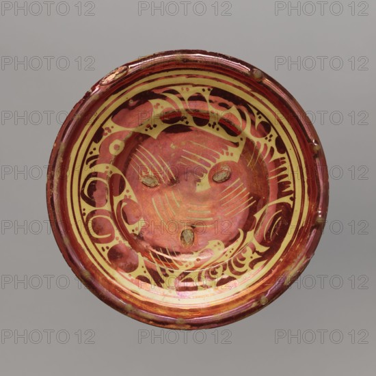 Plate, Manises ceramic center; ca. 1400-,  manufacturer author, mirrors decoration, birds decorative motif, plant decorations, Plate - a type of semi-deep bowl, with a thickened rim. Painted with red mirror in the motif of a bird with spread wings in the bottom, with volute plant motifs all around. Traces of supports used during firing on the face. 16th/17th century, Spain, Manises, vessel, height 4.5 cm, width 20.0 cm, A circular bowl with a glossy finish showing a pattern of swirling organic shapes and curved lines, featuring a central area with a few short, parallel incised strokes and three small oval depressions; the rim is slightly raised and shows minor irregularities. The visible colors include red, deep maroon, pale yellow, and hints of brown and greenish discoloration, with highlights that create a metallic sheen. The background behind the bowl is a flat, neutral gray.